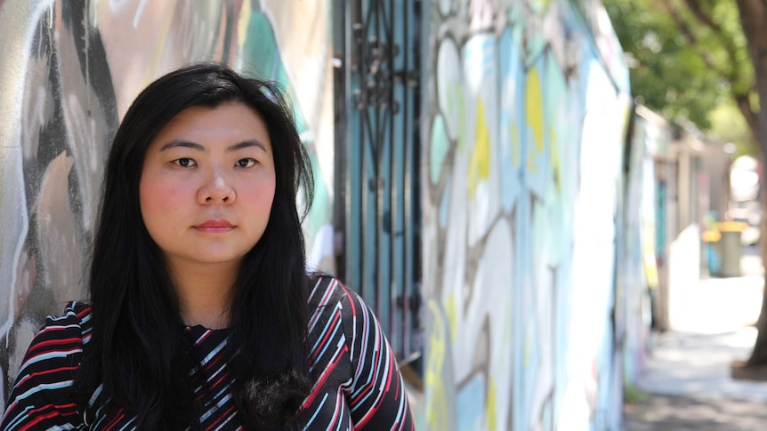 A woman leans against the bars outside a window, along a heavily graffitied wall with trees in the background.