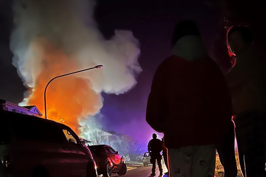 People stand on the street looking at an orange smoke cloud billowing from a house