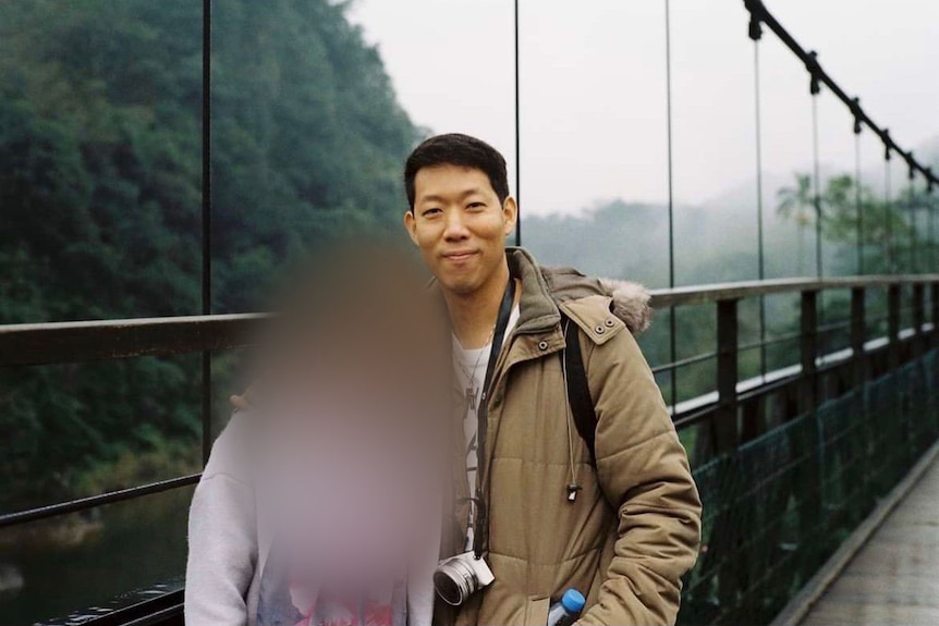A man with short dark hair poses for a photo on a bridge with another person whose face is blurred.