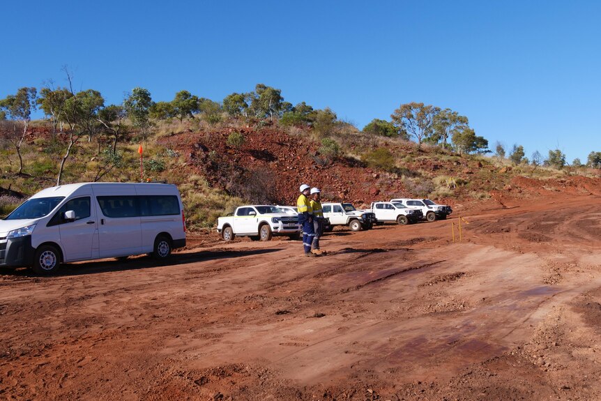 Two men in high vis jackets and white hard hats stand in front of a row of white utes, in front of a red hill.