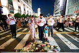 Three adults and a child lean over their hands and pray in front of flowers at a zebra crossing.