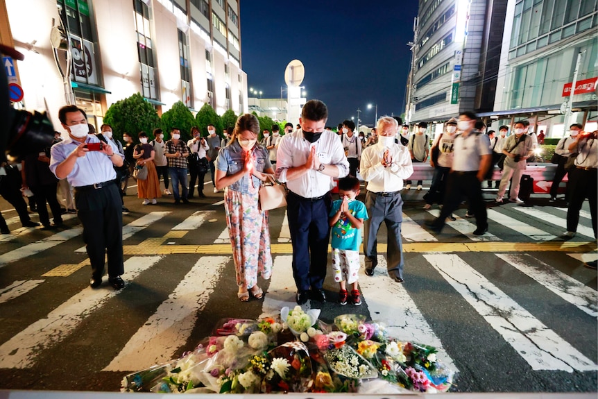 Three adults and a child lean over their hands and pray in front of flowers at a zebra crossing.
