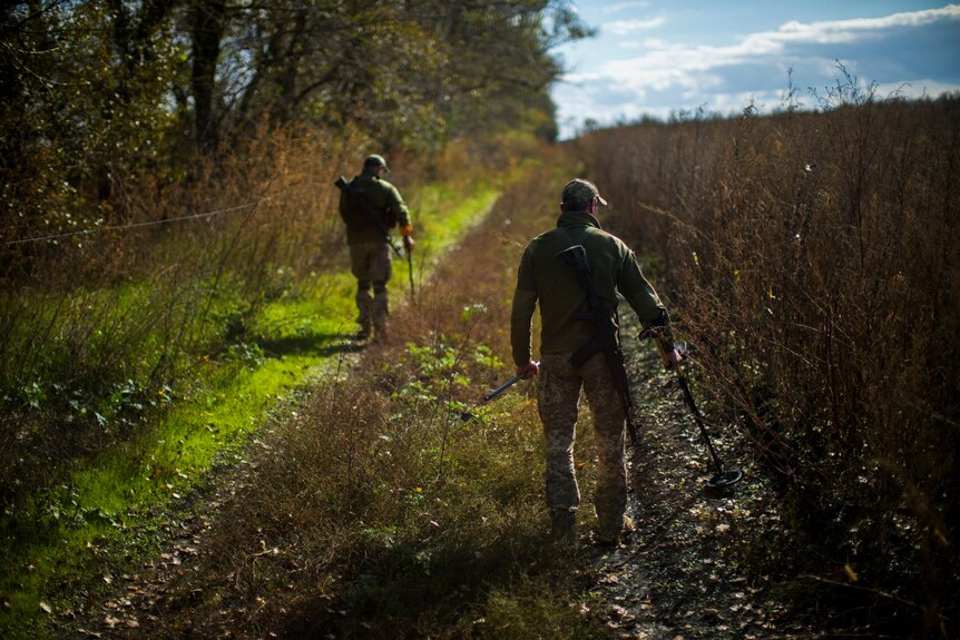 Two man in camouflage walk down a grassy trail with metal detectors in hand. 