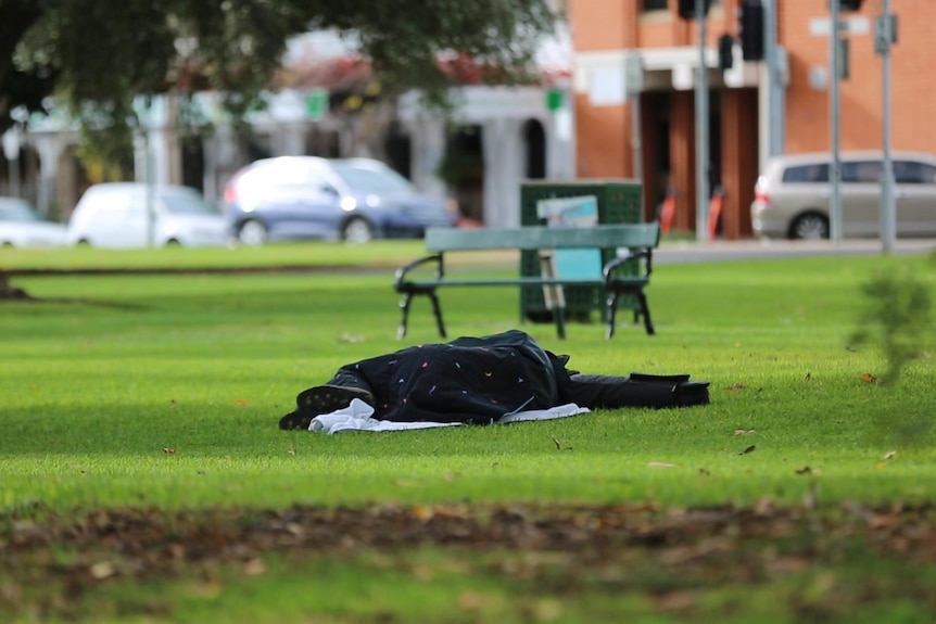 A person sleeping wrapped in a blanket in the middle of a grassy reserve, city streets and cars in the background