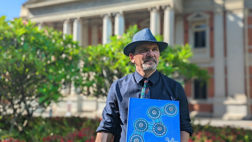 Eric Hayward stands in a garden at a historic court building with a blue dot painting by his missing sister Rebecca.