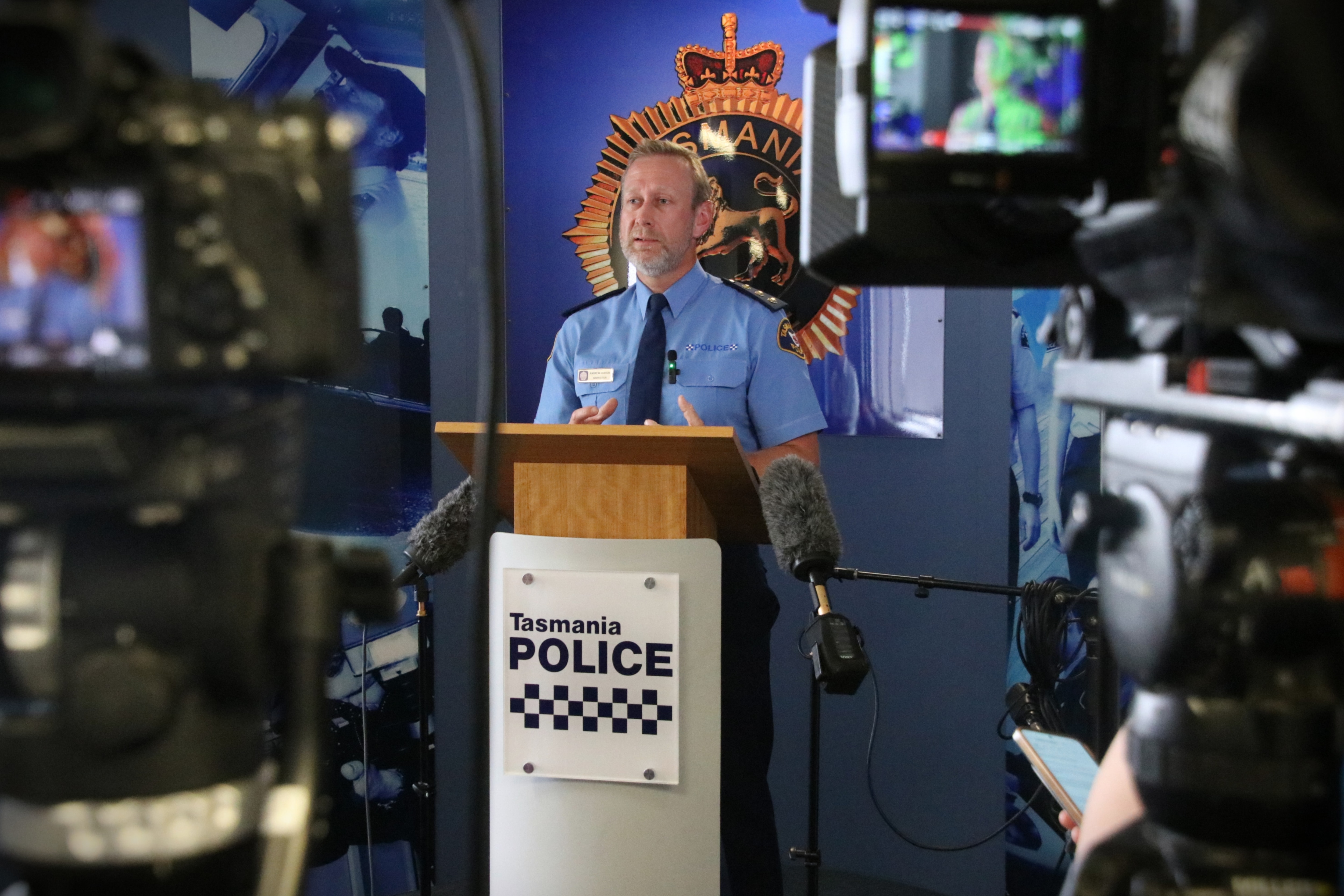 Um homem com uniforme policial azul marinho fica em um púlpito cercado por microfones, enquanto a mídia filma.