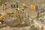Heavy machinery digs out a collapsed trench as emergency services personnel look on.