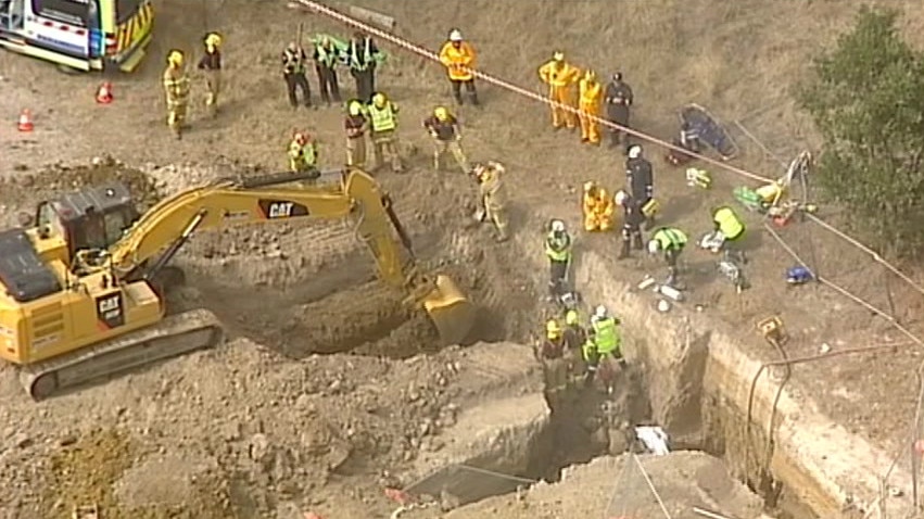 Heavy machinery digs out a collapsed trench as emergency services personnel look on.