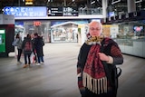 A woman with white hair wears a scarf at a train station with other people gathered behind her.
