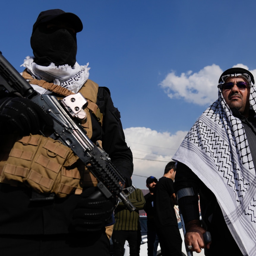 Two men, one armed with a gun, stand among a crowd of people at a funeral.