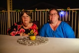 An older and a middle aged caucasian woman stare at the camera, sitting at a patio table in at night. The look serious.