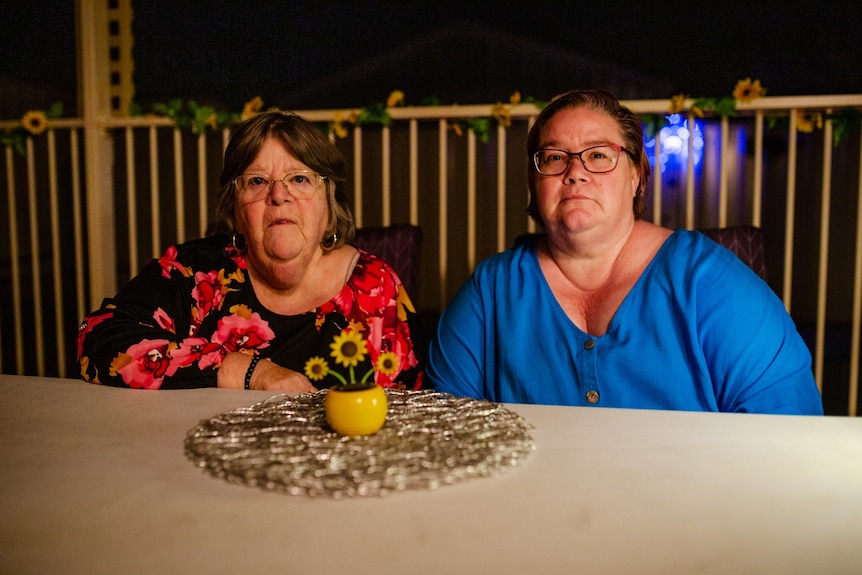 An older and a middle aged caucasian woman stare at the camera, sitting at a patio table in at night. The look serious.