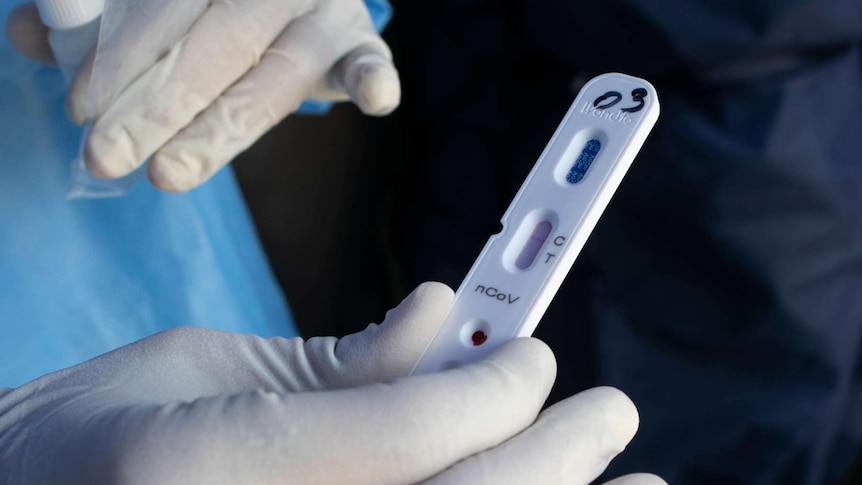 a person wearing white gloves and a blue gown holds up a coronavirus test