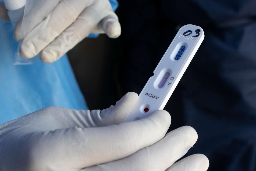 a person wearing white gloves and a blue gown holds up a coronavirus test