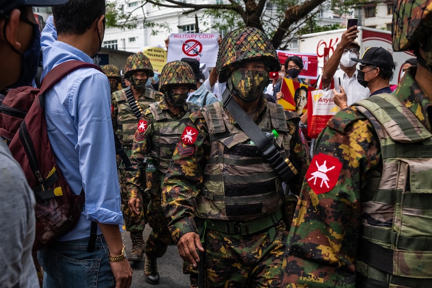 A line of soldiers wearing helmets and masks march through a crowd where people hold anti-coup signs.