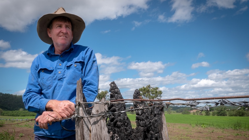 A man wearing an old hat leans on a barbed wire fence with a burnt fence post in the foreground.