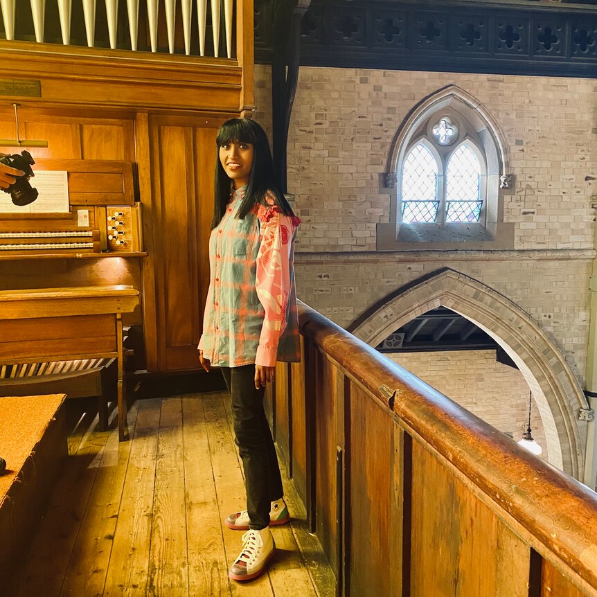 A young woman wearing a pink and blue shirt looks into the camera at a church with an organ in the background 