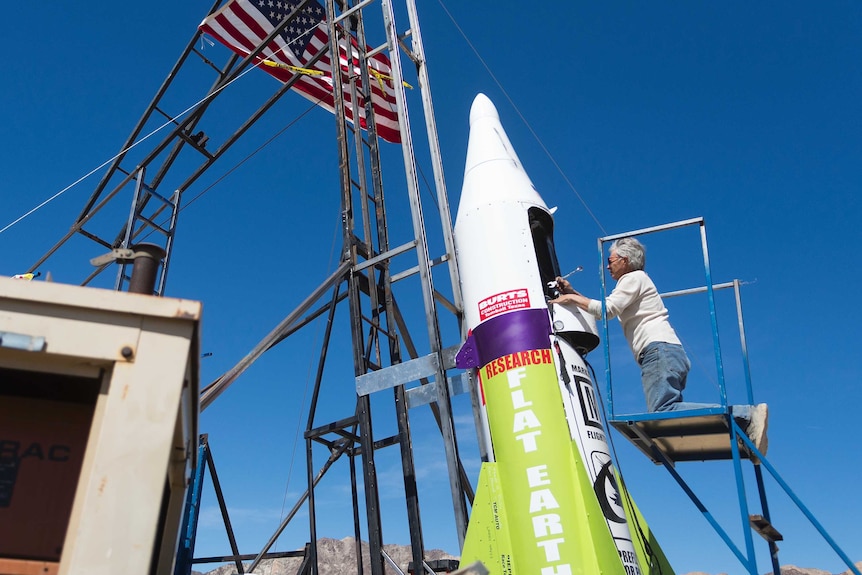 Man kneeling on a ladder repairing a home-made rocket.