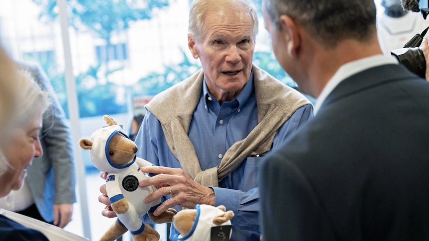 An older man wearing a chambray shirt & jumper tied around his shoulders, holds a spacesuit-wearing toy as he talks to locals.