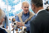 An older man wearing a chambray shirt & jumper tied around his shoulders, holds a spacesuit-wearing toy as he talks to locals.