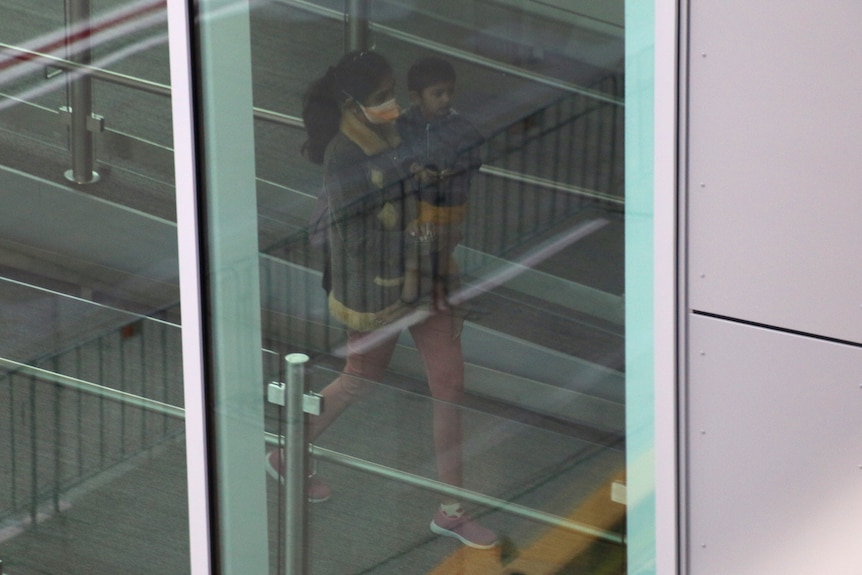 A woman walks along a corridor behind windows carrying a child and wearing a face mask.