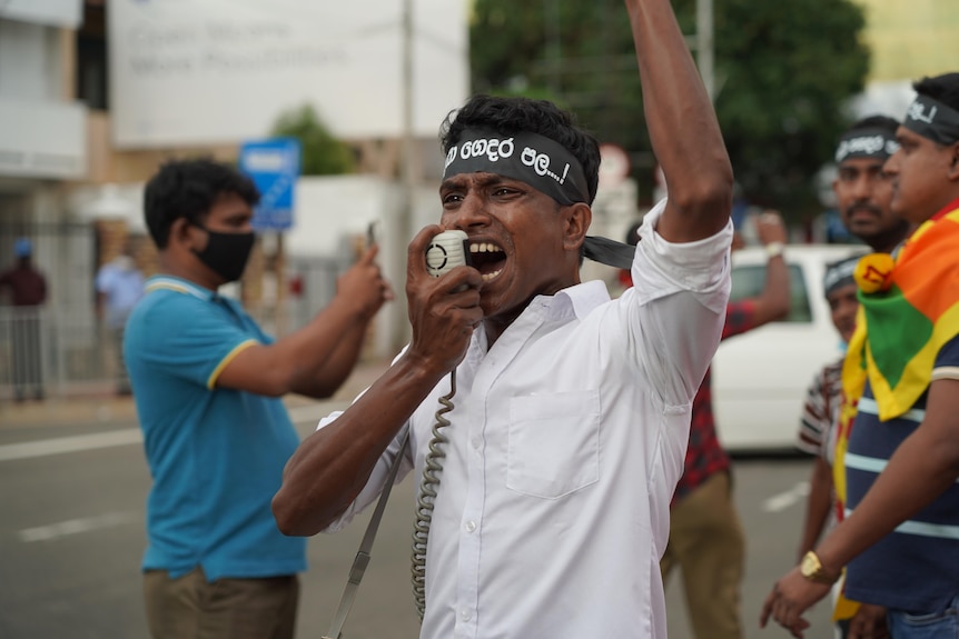 A man wearing a black band around his head shouts into the mouthpiece of a megaphone, his arm extended above his head