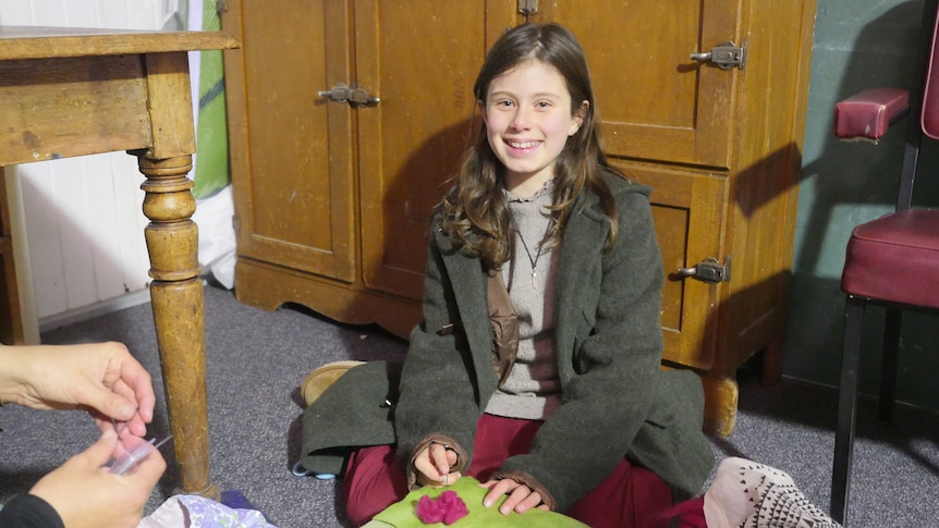 A young girl sits on the floor inside, she is surrounded by different fabric