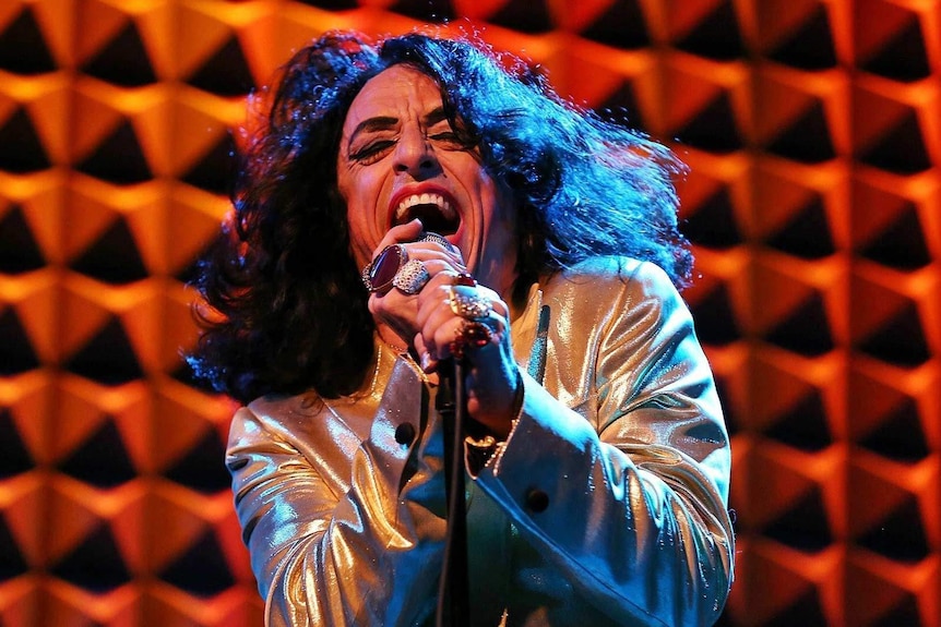 The singer Paul Capsis in silver shirt and long hair, belting into a microphone, orange wall in the background