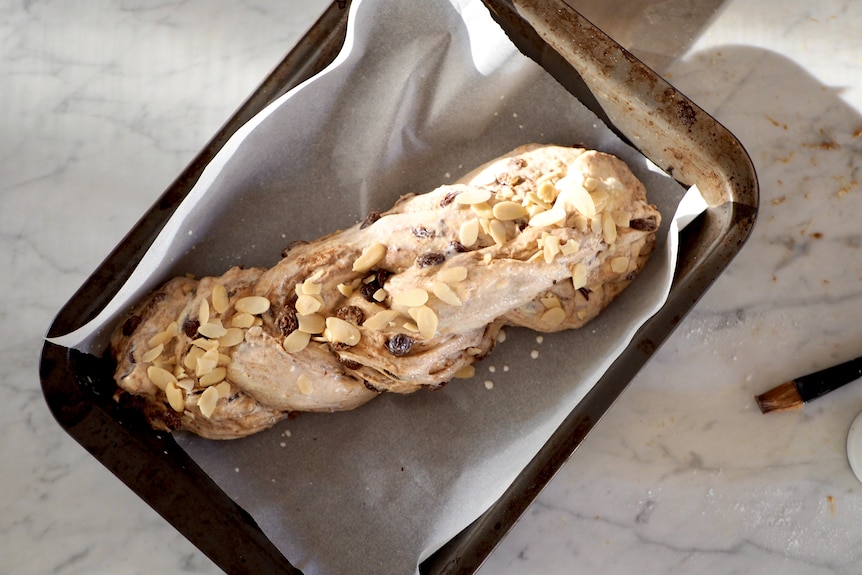 A raw loaf of bread, knotted together and dusted with almonds, sitting on baking paper in a tray.