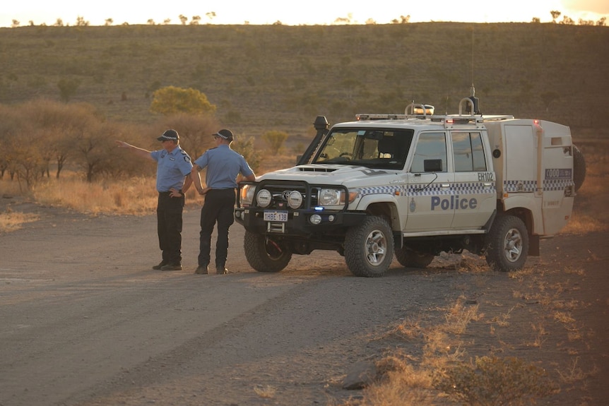 Two men in uniform stand on a dusty road near a police car