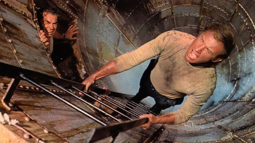 A still from the film The Poseidon Adventure, with a man climbing down a ladder into a metal shaft, with a pained expression