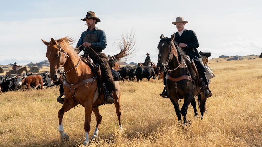 A scruffy 40-something man and a more polished 30-something man, each wearing a cowboy hat, ride horses through a field