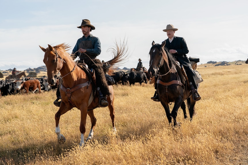 A scruffy 40-something man and a more polished 30-something man, each wearing a cowboy hat, ride horses through a field