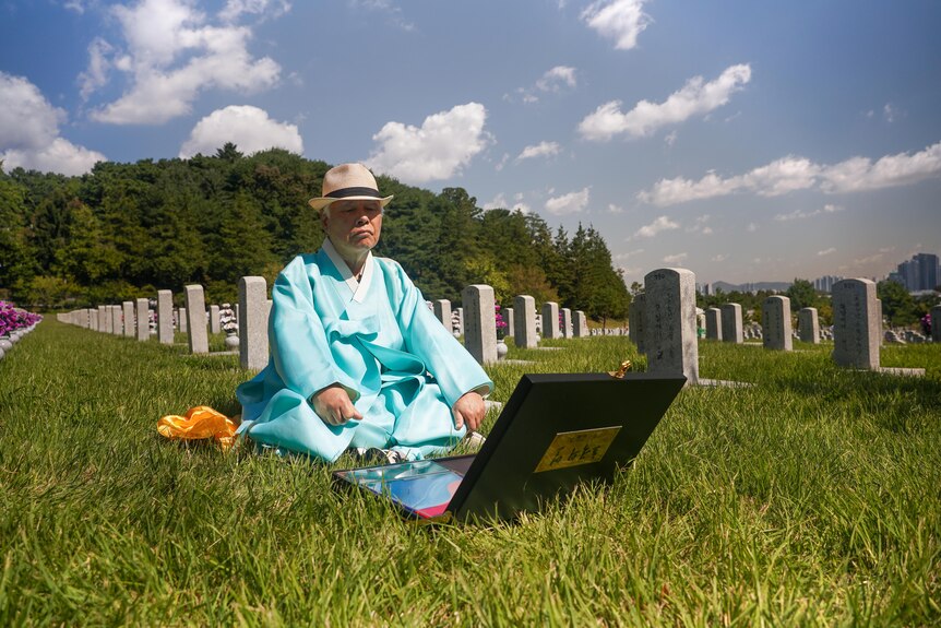 A man wearing blue robes and a hat sits on lawn in front of a box and surrounded by gravestones