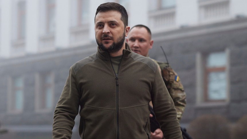Volodymyr Zelenskyy walks outside wearing a khaki zip up jacket. Behind him is a solider in camo. 