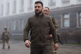 Volodymyr Zelenskyy walks outside wearing a khaki zip up jacket. Behind him is a solider in camo. 