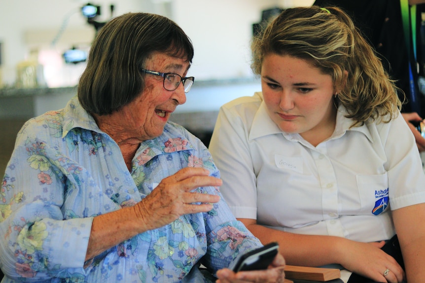 An retired woman  wearing a blue floral top talks to a teenage girl in school uniform who is listening intently
