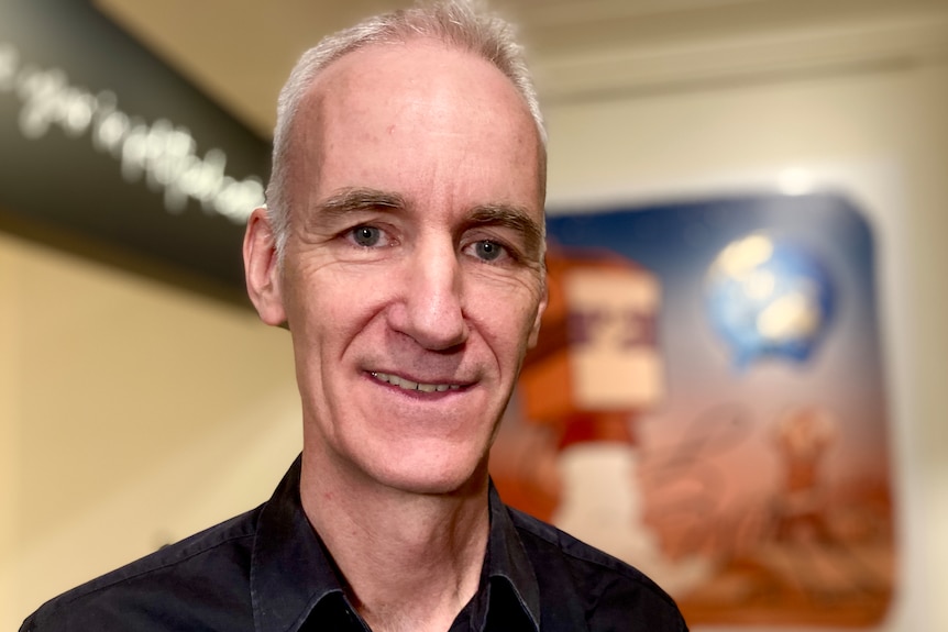 A close up photo of a middle aged man smiling, with a dark shirt against a blurred background. 