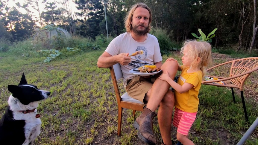 A person eating chicken while being watched by a small girl and a dog.