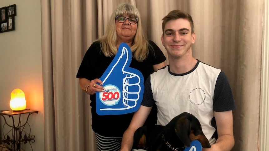 Mum with blonde hair holding giant blue thumbs up standing next to boy in wheelchair