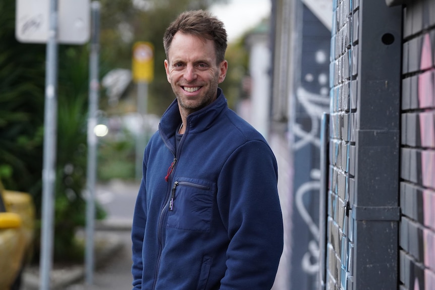 A man in a navy sweater stands in front of a graffiti wall smiling