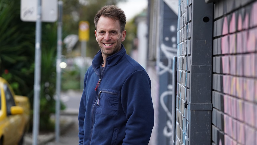 A man in a navy sweater stands in front of a graffiti wall smiling