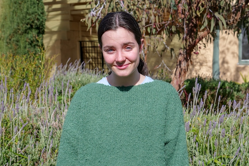 A girl with dark hair wearing a green jumper looks at the camera.