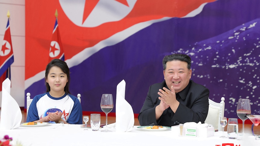 A man in black suit sits with a girl in front of North Korea flag.