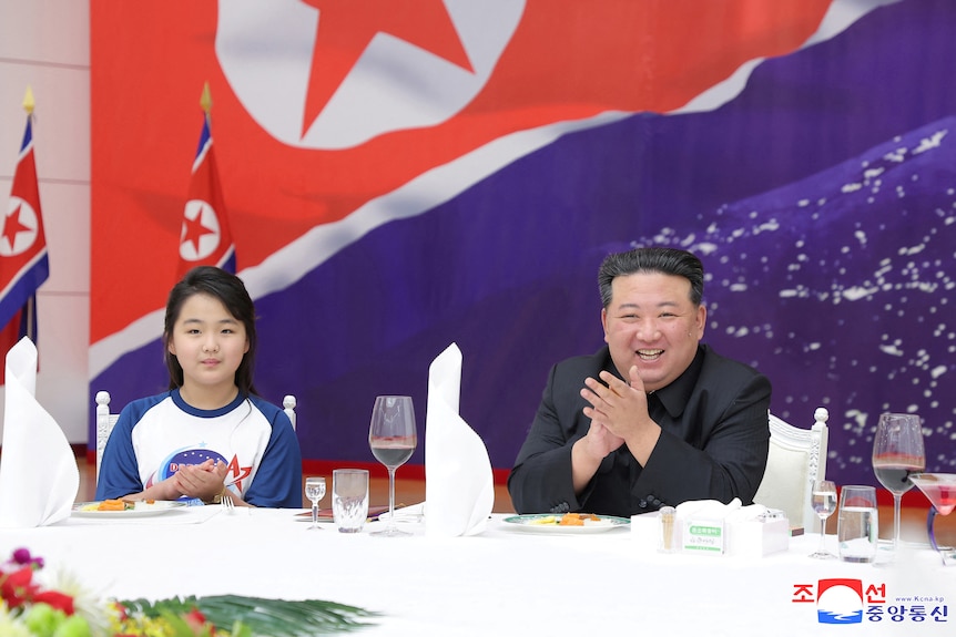 A man in black suit sits with a girl in front of North Korea flag.
