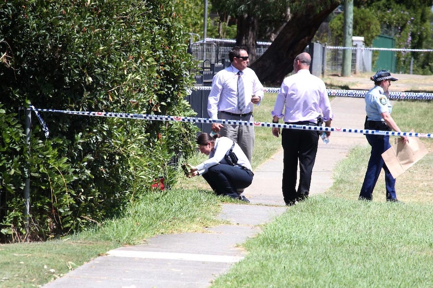 A plain clothed police officer crouches down, photographing a red container in a bush, next to other police.