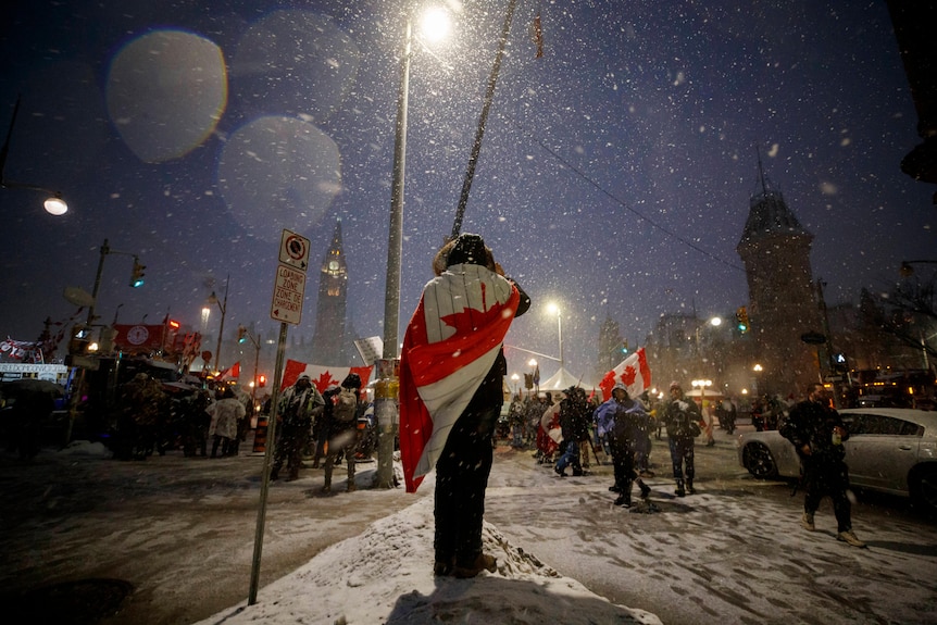 A protester stands in the snow, drapped in a Canada flag, with several more protesters in the backgrounds.