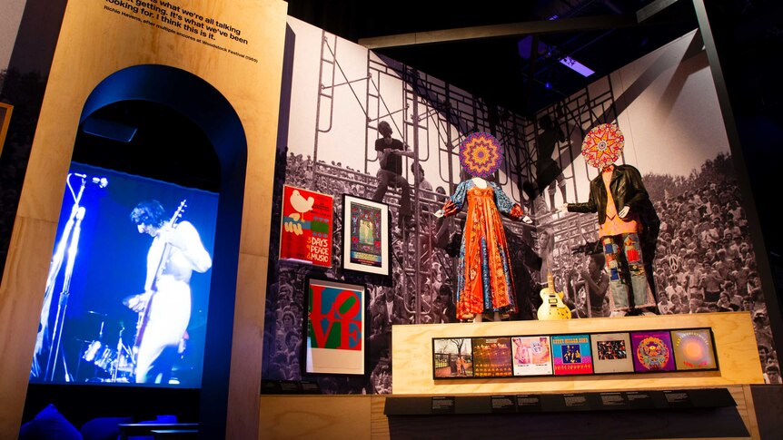 Exhibition view: A video of a rock band through a doorway, mannequins in colourful 60s clothes and psychedelic posters.