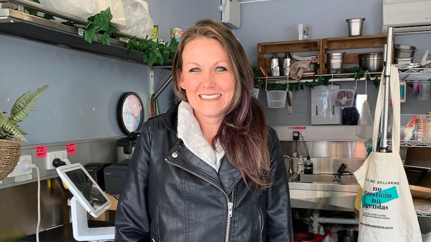 A woman with long brown hair and a black leather jacket stands in a cafe kitchen.