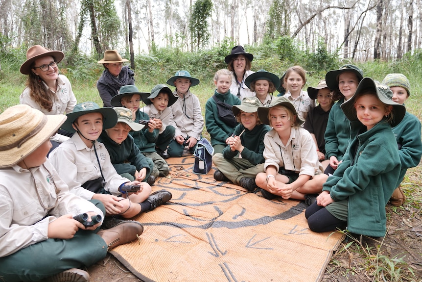 A group of primary school students wearing green hats and khaki shirts sit on a mat in a bush setting with their principal